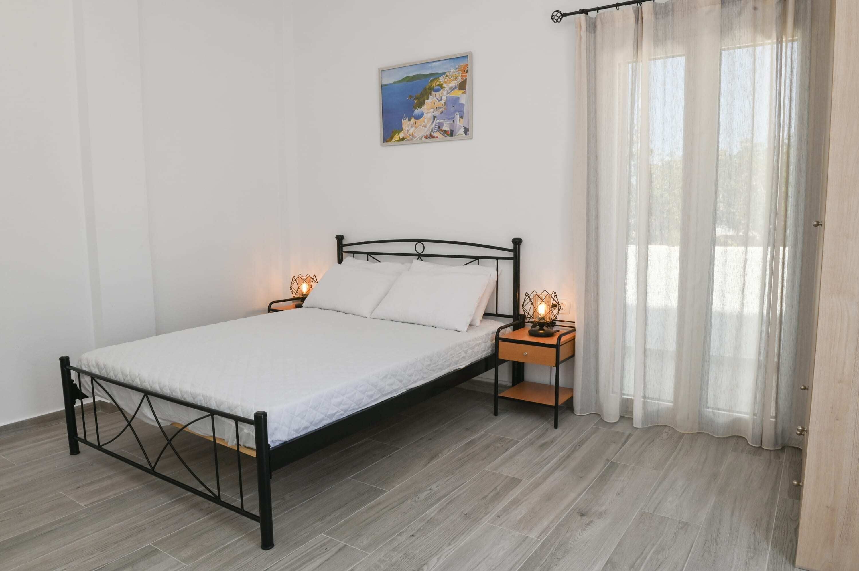 Un dormitorio con una cama individual, una mesita de noche, una lámpara de noche, un cuadro en la pared y una ventana con cortinas translúcidas.