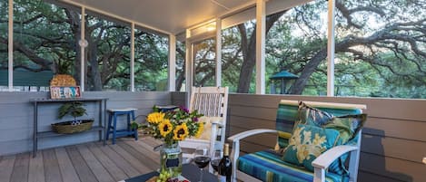 A covered outdoor seating area with a wooden table set with food, a bottle of wine, and a vase of sunflowers.