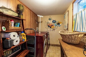 A laundry room with a washing machine, shelves with cleaning supplies, a decorative fish mural, and a window with curtains. - Red Bird Cabin - Studio Retreat (Kingsland)