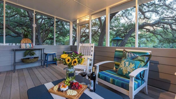 A covered outdoor seating area with a wooden deck, a white chair with a blue cushion, a table with a fruit platter, a bottle of wine, and a vase with sunflowers. - Red Bird Cabin - Studio Retreat (Kingsland)