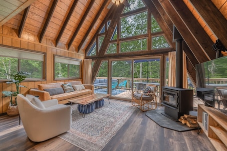 A spacious living area with a high wooden ceiling, large windows, a fireplace, and a television.