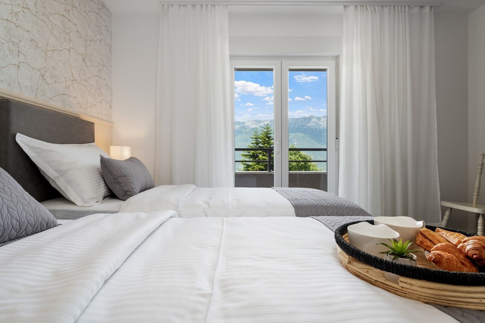 Ein Hotelzimmer mit zwei Betten, Blick auf Berge durch das Fenster und einem Frühstücksbrett mit Croissants.