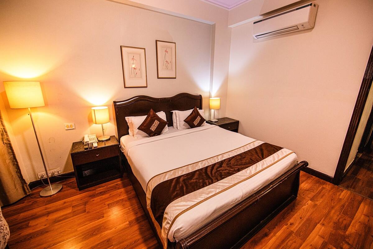 A neatly made bed with a wooden headboard, two bedside lamps, and a wall-mounted air conditioner.