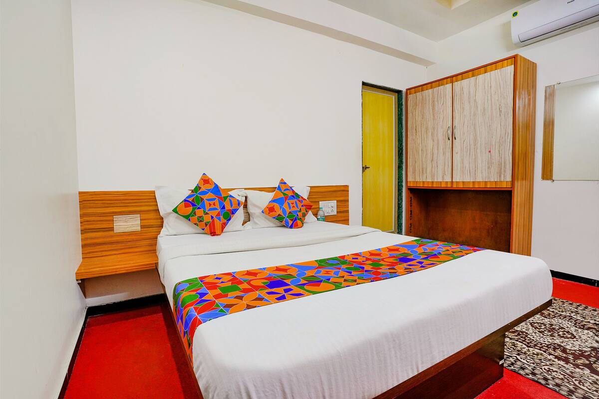Cama bem arrumada com colcha colorida estampada com padrões geométricos, cabeceira de madeira e tapete vermelho que leva à porta.