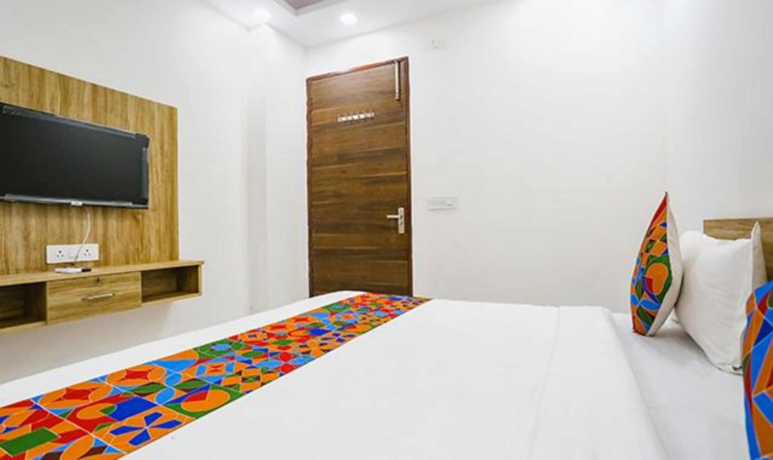 Ein Hotelzimmer mit Bett, Wandfernseher, Holztür und bunter Tagesdecke.