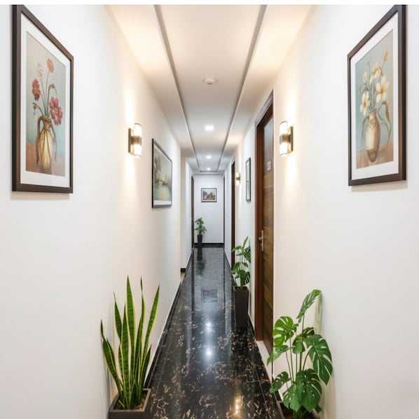 Un couloir aux murs ornés de tableaux floraux encadrés, doté d’un sol noir poli et bordé de chaque côté de plantes en pot.