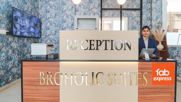 A reception desk with a sign reading "RECEPTION" and "BROHOLIC SUITES" below it. There is a television mounted on the wall above the desk displaying images. A person is seated behind the desk, and a laptop is on the desk. - FabHotel Broholic Suites I (Hyderabad)