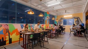 A dining area with tables and chairs, large windows, and colorful wall art. - FabHotel Broholic Suites I (Hyderabad)