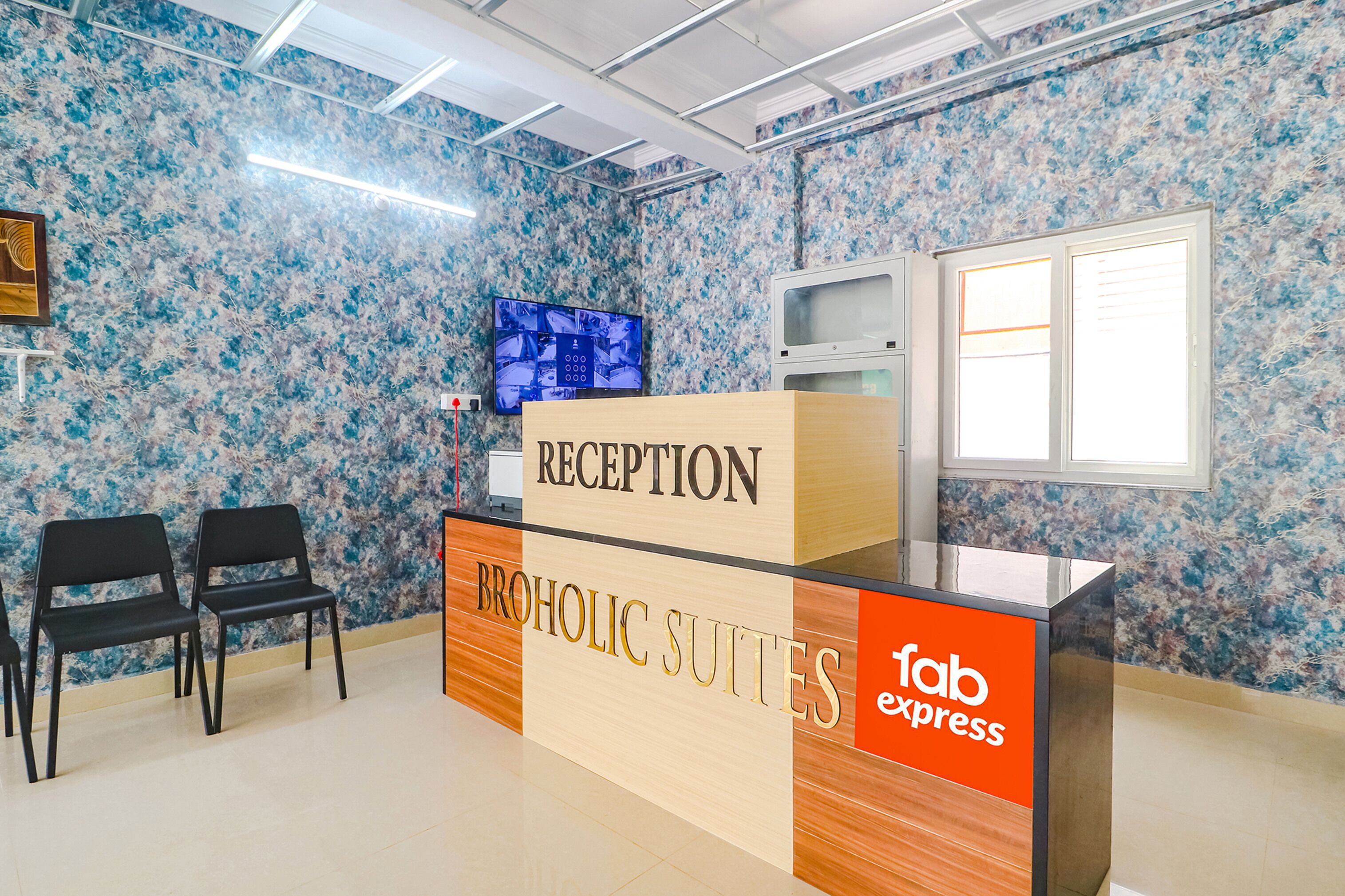 Un comptoir d’accueil portant les inscriptions « RÉCEPTION » et « BROHOLIC SUITES », avec une pancarte « fab express », deux chaises noires, un téléviseur et un four à micro-ondes fixés au mur.