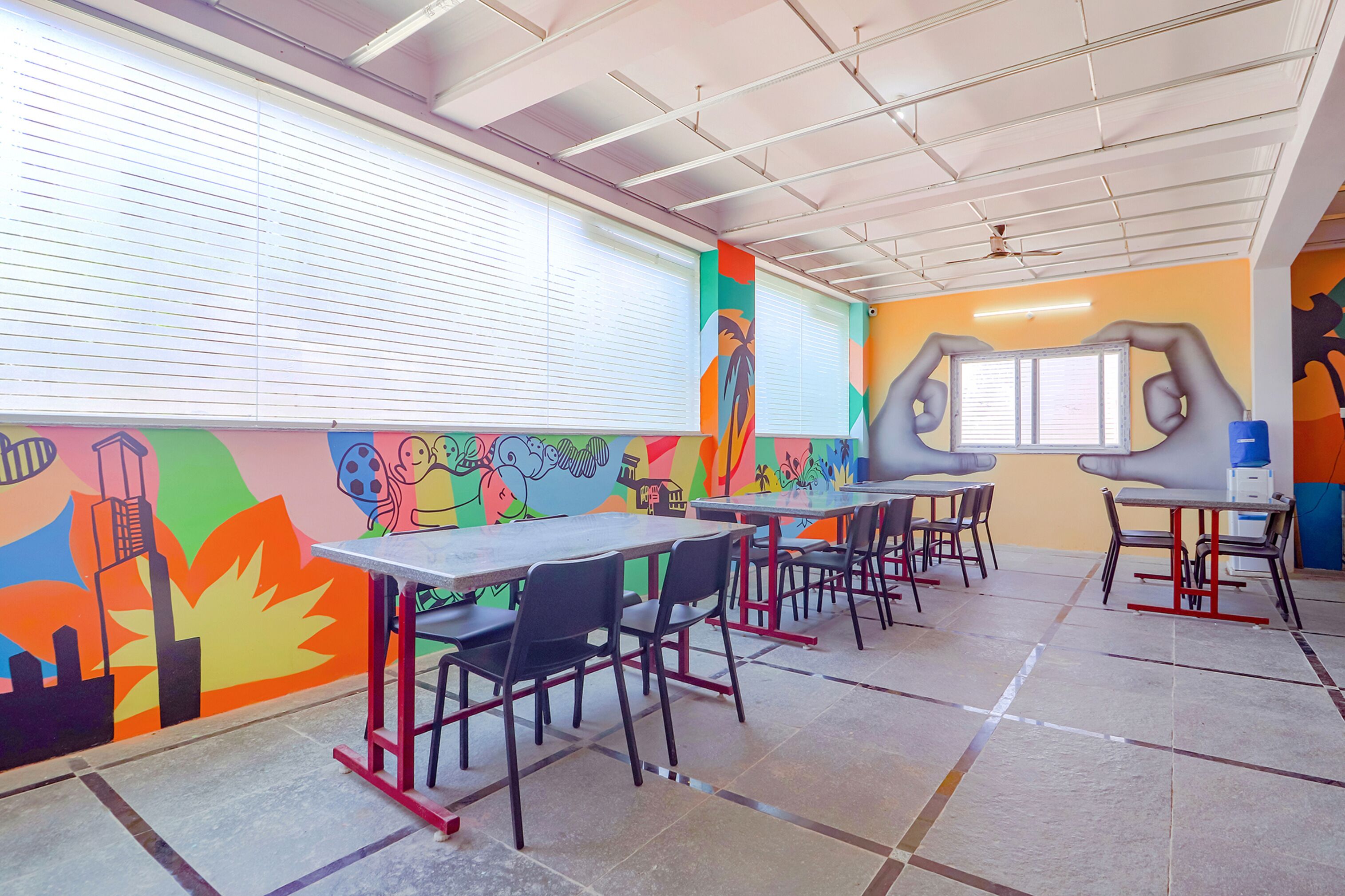A cafeteria with tables and chairs, colorful murals on the walls, and a ceiling fan.