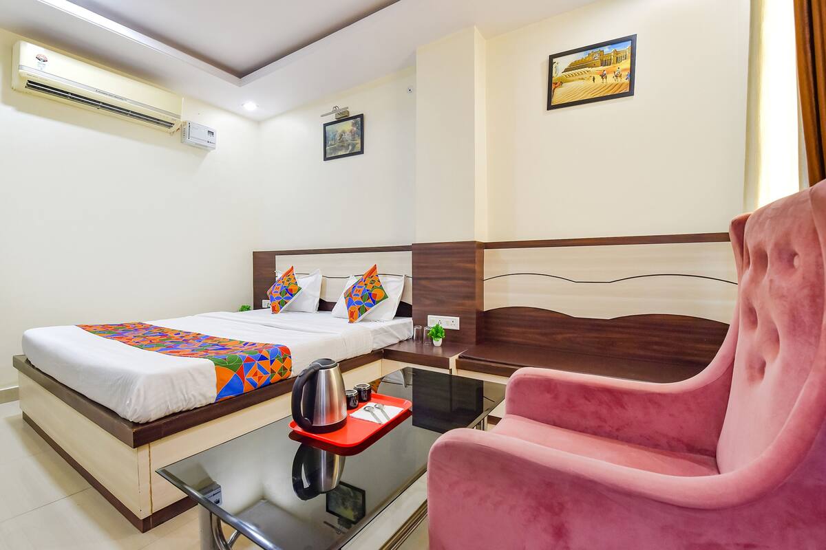 A hotel room with a bed, a red tray with a kettle, a pink chair, and two paintings on the wall.