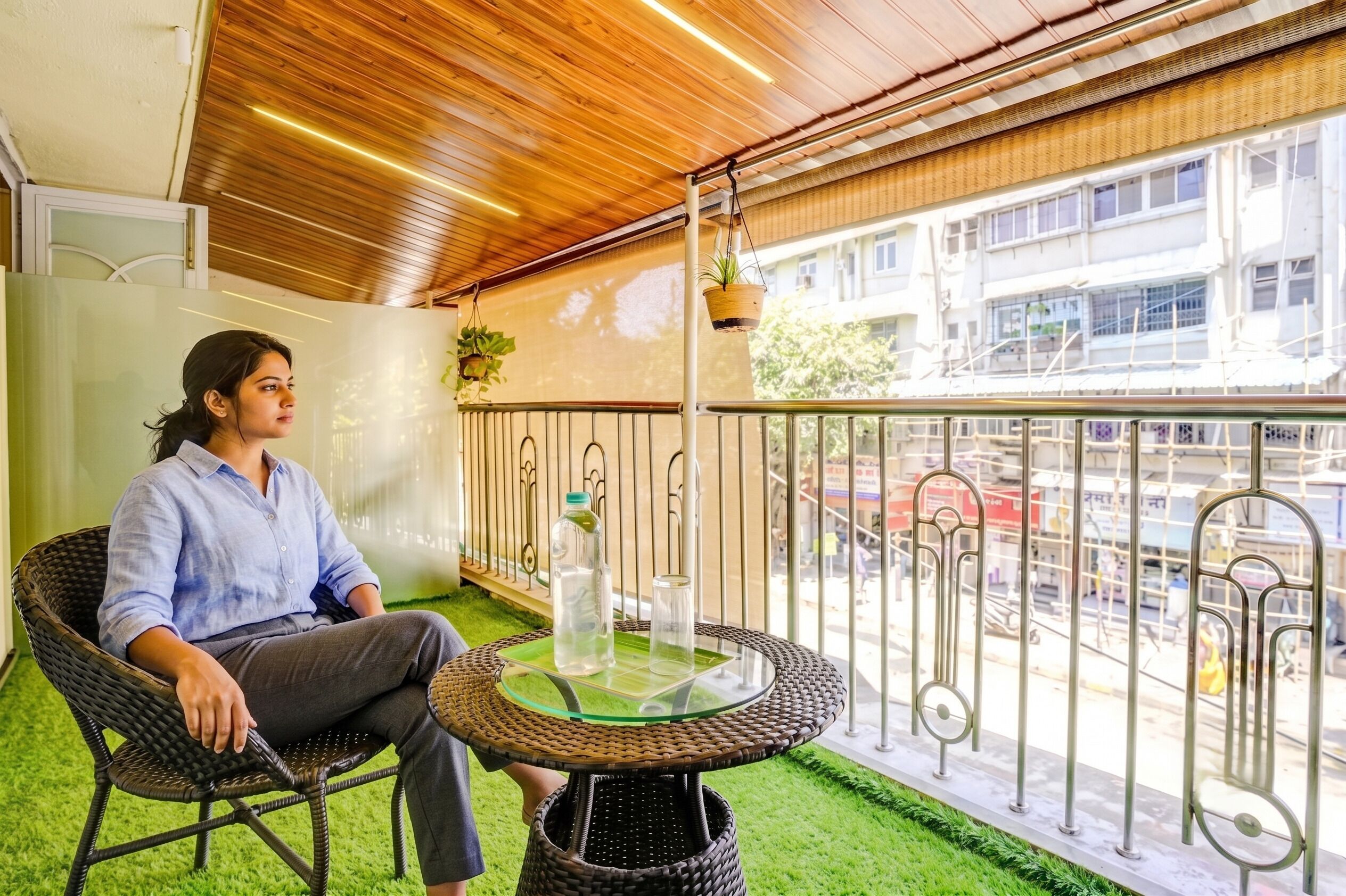 Une personne assise sur une chaise en osier, sur un balcon, devant une table en verre, avec une plante suspendue, et une vue sur une rue et des immeubles.