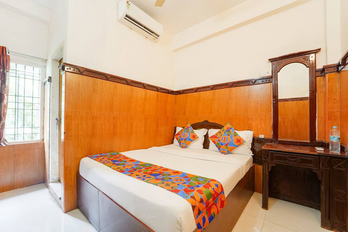 A hotel room with a wooden headboard, a bed with colorful bedding, a bedside table with a bottle, and a mirror.