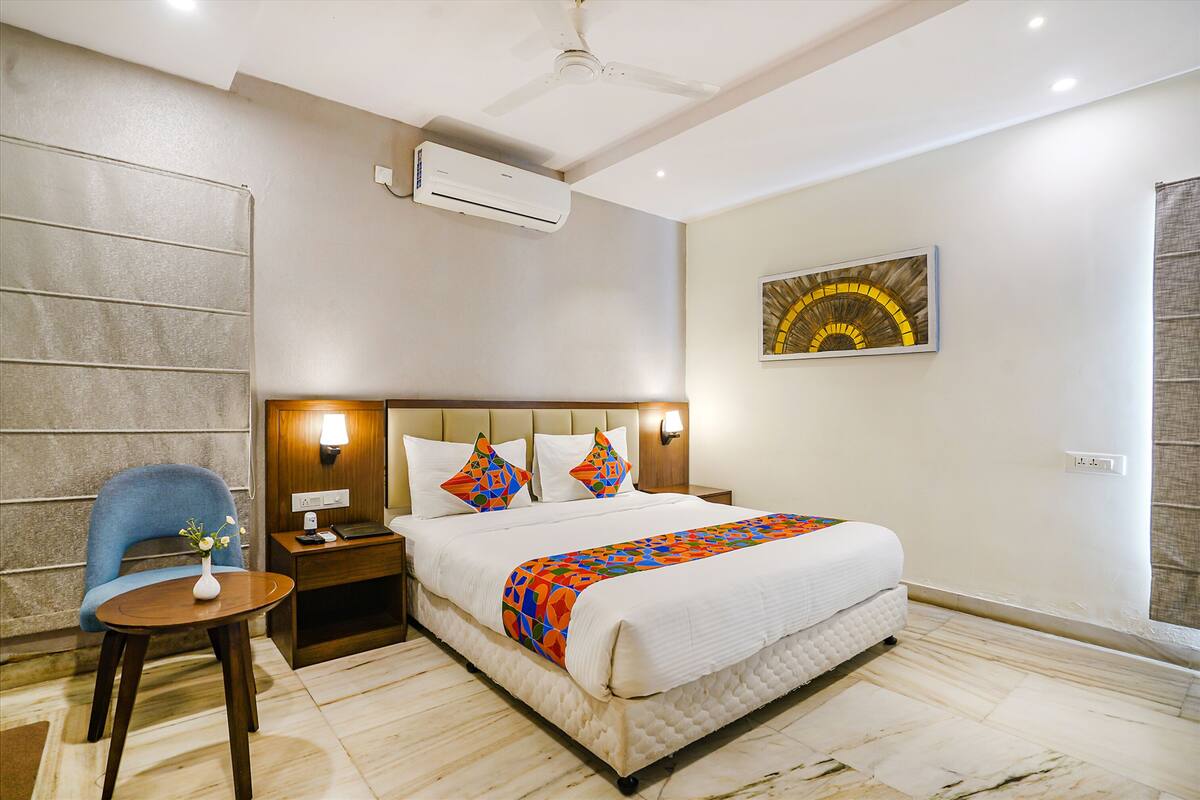 Habitación de hotel con cama, una silla, una mesita pequeña, aire acondicionado y una lámpara de pared.