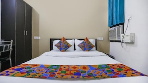 A hotel room with a bed, two pillows with colorful geometric patterns, a white bedspread, a colorful geometric-patterned throw blanket, a dark wardrobe, a window with a blue curtain, and an air conditioning unit on the wall.