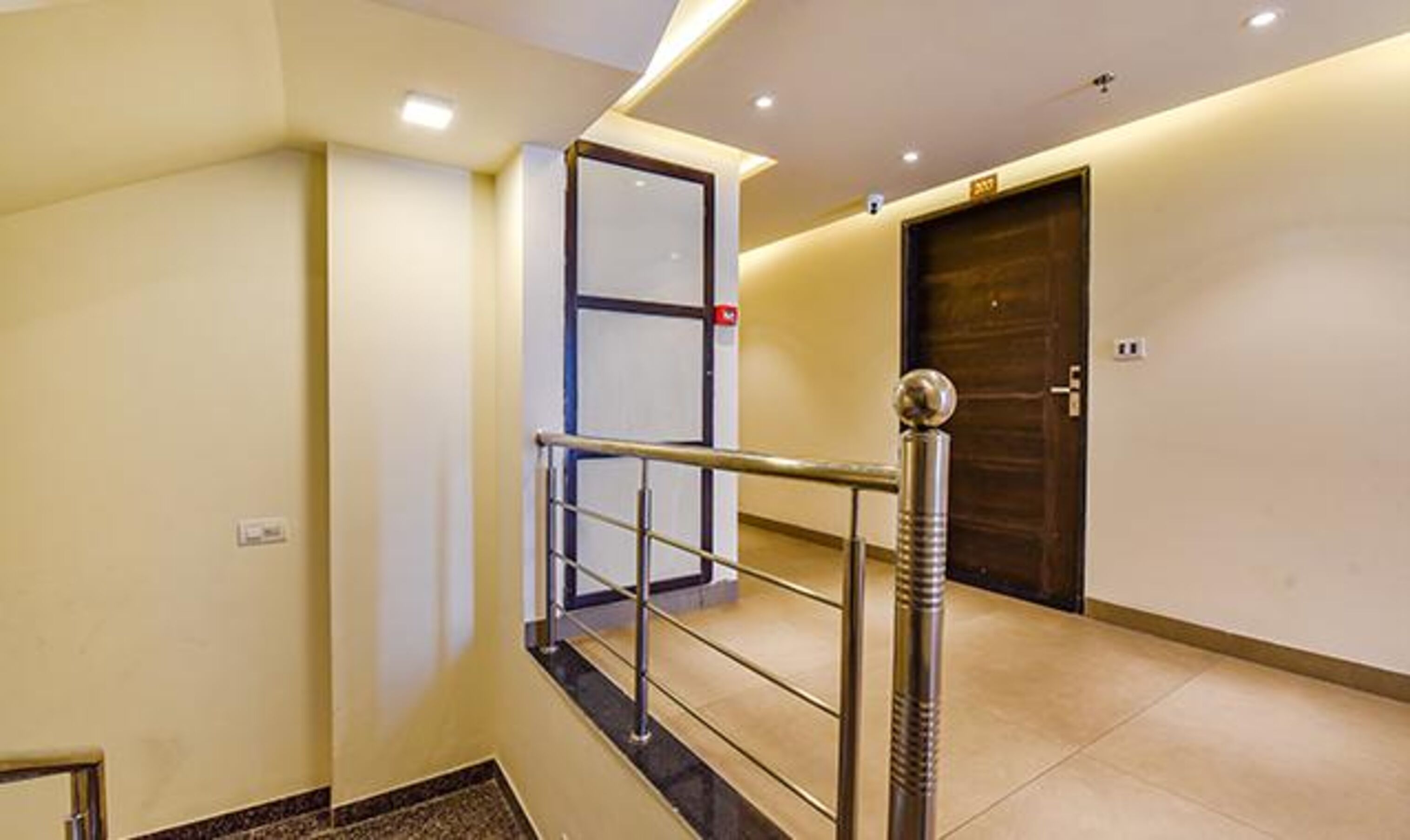 A modern hotel corridor with a metal railing, a wooden door, and a glass elevator door.