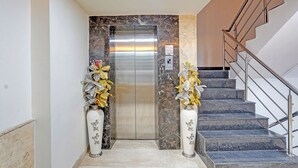 Un ascenseur avec un mur en marbre, un escalier avec des rampes en métal et deux vases décoratifs contenant des fleurs artificielles.