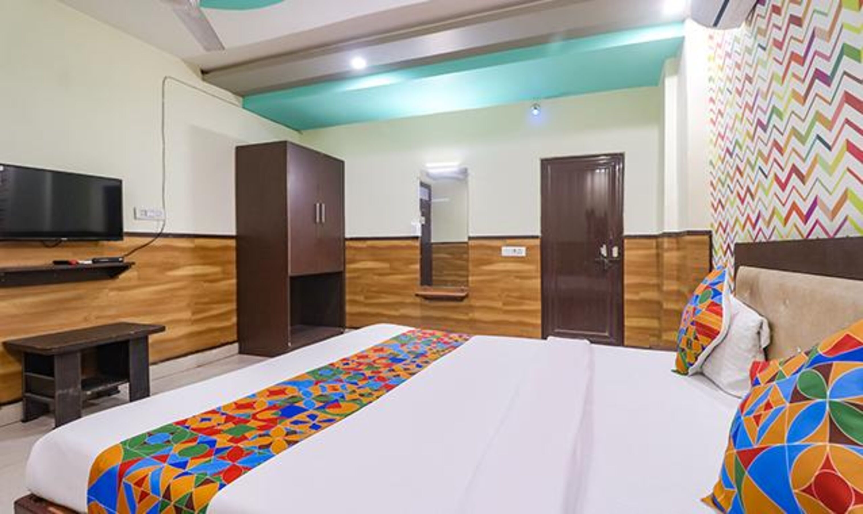 A hotel room with a large bed, a TV, wooden paneling, a mirror, and a colorful wall design.