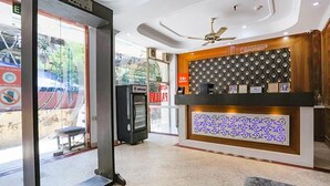 A reception area with a modern design, including a patterned wall, a ceiling fan, and a counter with framed items. - Fabexpress Station Ii (New Delhi)
