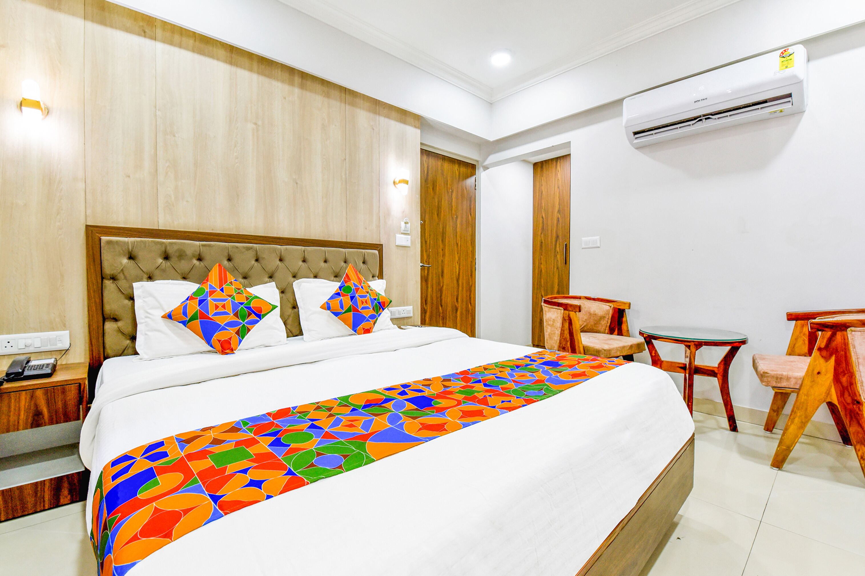 A hotel room with a large bed, a wooden headboard, a colorful bedspread, a small table, and a chair.