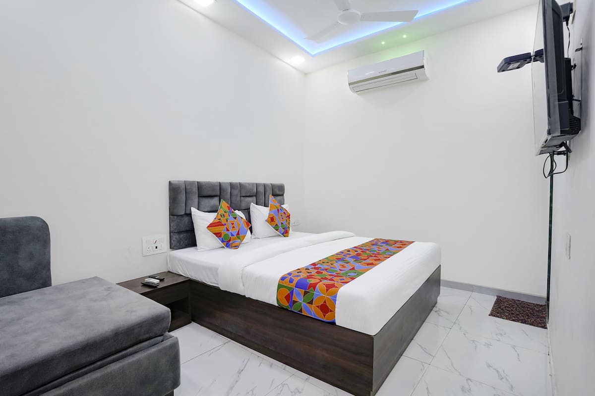 A modern hotel room with a bed, a grey sofa, a TV mounted on the wall, and an air conditioning unit.