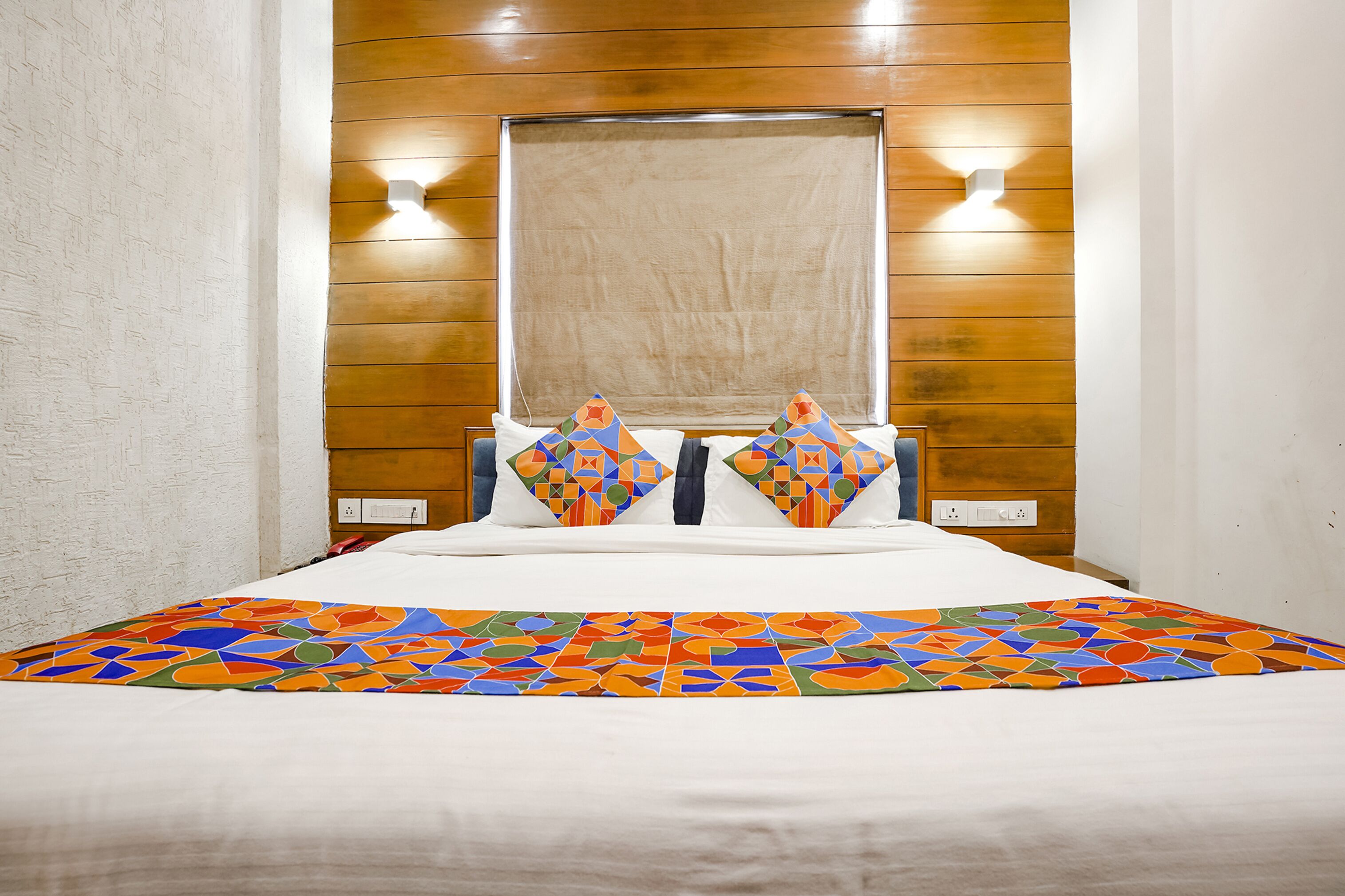 Hotelkamer met een houten hoofdbord, twee wandlampen, een groot raam en een kleurrijke deken met een geometrisch patroon.