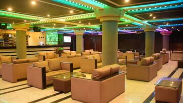 Un hall con sofás y sillones dispuestos alrededor de pilares, una zona de bar con estanterías y una pantalla mostrando contenido.