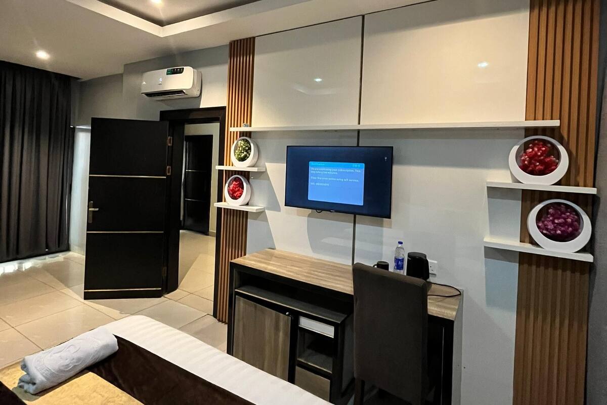 Une chambre d’hôtel moderne équipée d’un lit, d’un bureau, d’une chaise, d’une télévision et d’étagères intégrées où sont disposés des objets décoratifs.