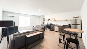 A modern living room with a grey sofa, a wooden coffee table, and a kitchen area with a stainless steel refrigerator, stove, and microwave.