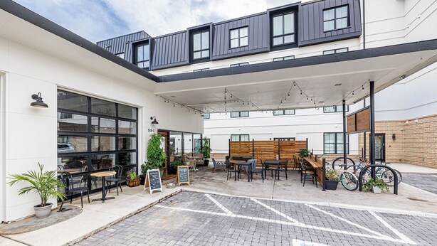 A modern building with a covered outdoor seating area, large windows, and a brick wall.