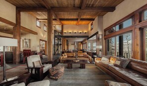 A spacious living room with a high ceiling, wooden beams, and large windows offering a view of the outdoors. - Elevated Mountain Ski Home by InvitedHome | New Remodel, Patio Views, Game Room (Mountain Village)