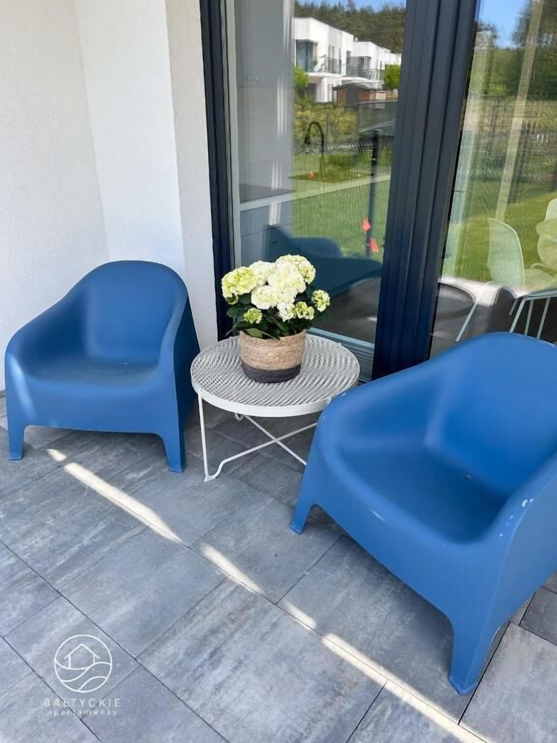 Un balcón con dos sillas de plástico azul, una pequeña mesa redonda con una cesta trenzada que contiene flores blancas, y una puerta de cristal que da a un jardín.
