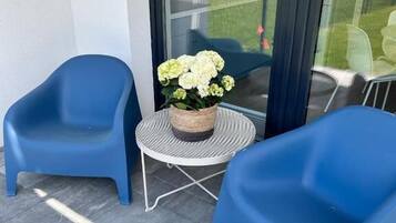 Un balcón con dos sillas de plástico azul, una pequeña mesa redonda con una cesta trenzada que contiene flores blancas, y una puerta de cristal que da a un jardín.
