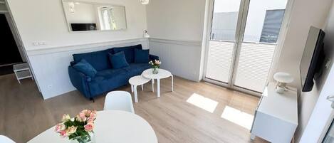 A modern living room with a blue sofa, a white dining table, and a small white table. There is a vase with flowers on the dining table.