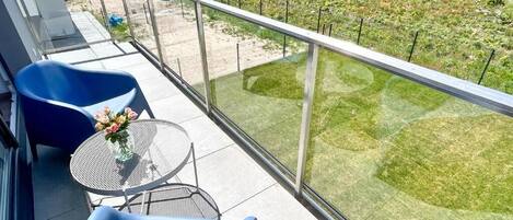 A balcony with blue chairs, a small round table with a vase of flowers, and a view of a well-maintained garden.