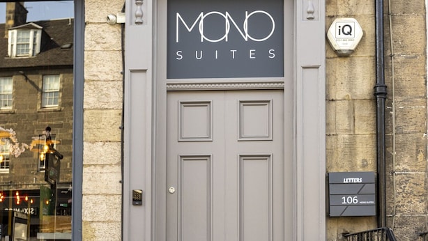 L’entrée d’un immeuble se compose d’une double porte grise, d’une enseigne indiquant « MOND SUITES » et de deux plantes en pot placées sur les marches.
