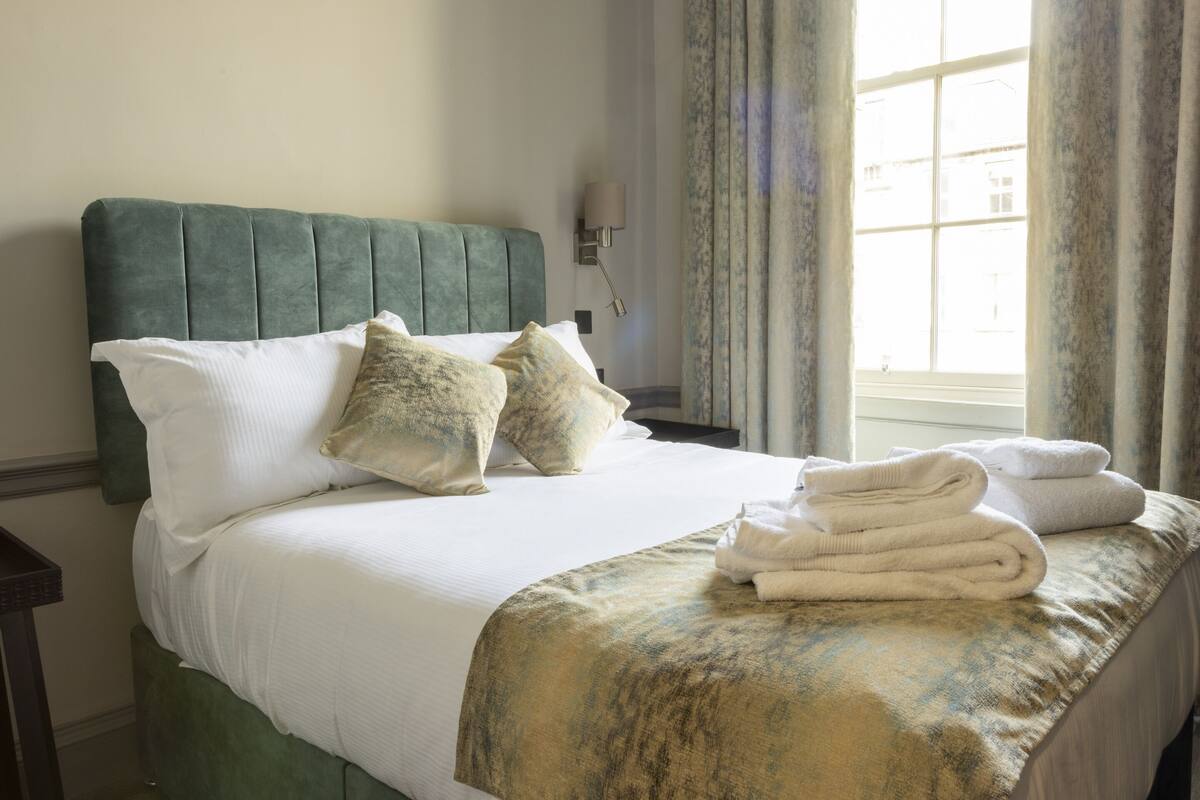 A neatly made bed with a green headboard, white linens, and a folded towel on the side.