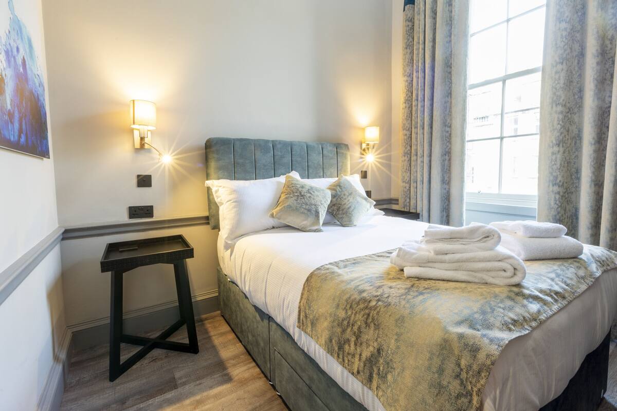 A neatly made bed with white linens and a blue and yellow patterned blanket, two bedside lamps, a small black side table, and a window with curtains.