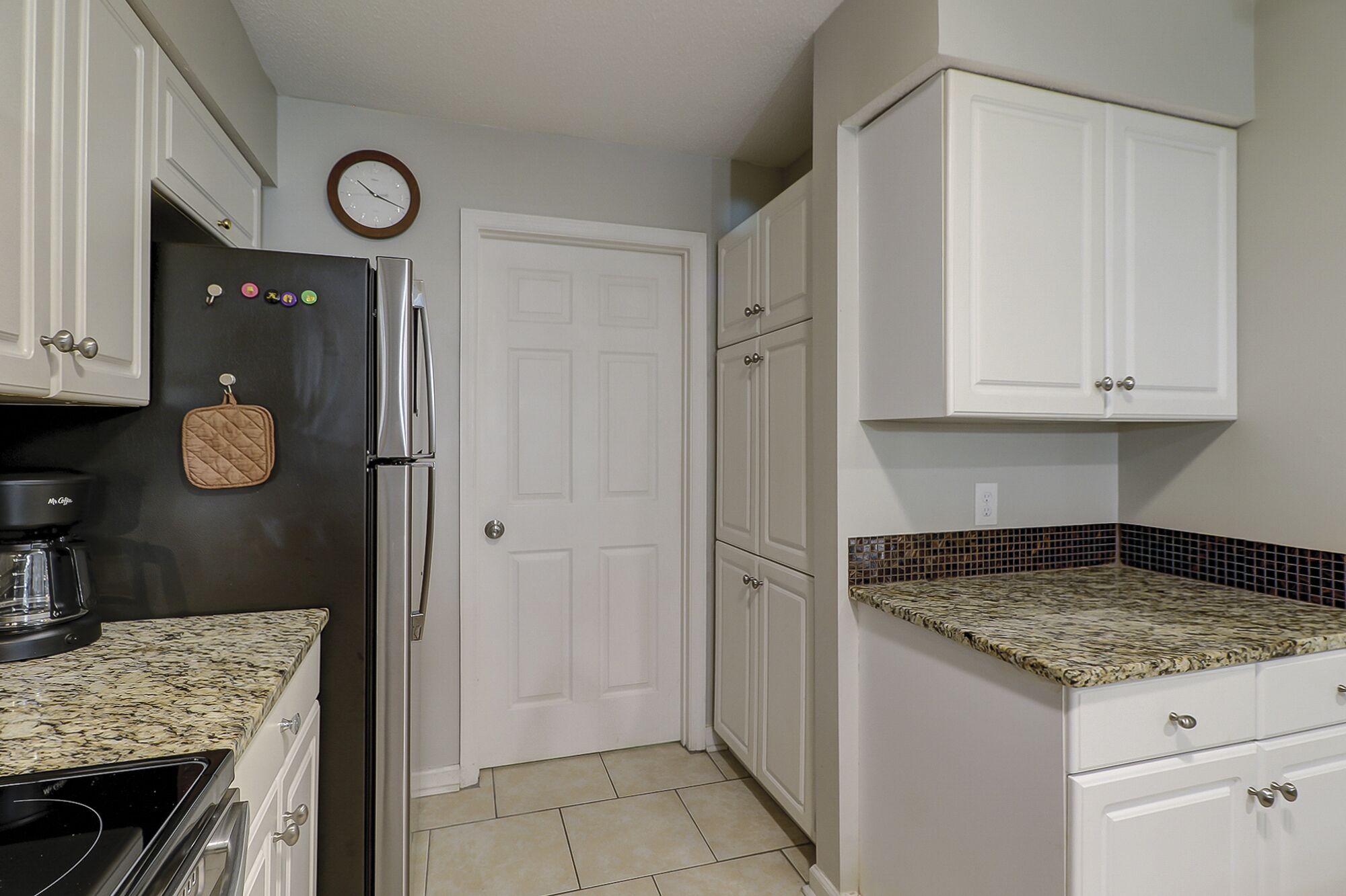 A kitchen with white cabinets, a black refrigerator, a coffee maker, and a clock on the wall.