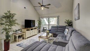 A living room with a grey sofa, a TV on a wooden stand, a ceiling fan, and a large window with a view of greenery. - Sunset Rentals | Colonnade Club 142 (Hilton Head Island)