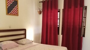 A bedroom with a bed, a wall-mounted picture, red curtains, and a window with blinds. - Magic Building Apartment 4 (Tarrafal)