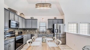 A modern kitchen with grey cabinets, stainless steel appliances, a wooden dining table, and a chandelier. - Trinity Tides, the waves dance, and so do your worries (Port Aransas)