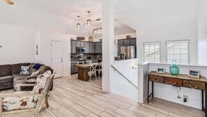 A spacious living area with a sofa, coffee table, and a dining table with chairs. A kitchen with modern appliances is visible in the background. - Trinity Tides, the waves dance, and so do your worries (Port Aransas)