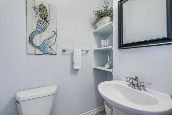 A bathroom with a white toilet, a sink with a mirror, and a decorative mermaid painting on the wall. - Trinity Tides, the waves dance, and so do your worries (Port Aransas)