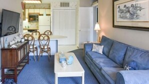 Una sala de estar con un sofá azul, una mesa de centro blanca y un mueble de madera. Hay un televisor a la izquierda y un cuadro enmarcado en la pared.