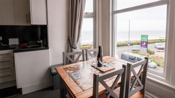 Une petite pièce comprenant une table à manger, des chaises, une fenêtre donnant sur la mer et un coin cuisine avec des placards et un évier.