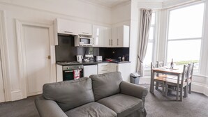 Un salon moderne avec un canapé gris, une table à manger avec des chaises, un coin cuisine doté de meubles blancs, un four à micro-ondes et une fenêtre avec des rideaux.