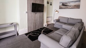 Un salon moderne avec un canapé gris, un téléviseur à écran plat fixé sur un panneau en bois, un lampadaire et un tapis noir et blanc.