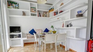 Een gezellige, compacte leefruimte met een eethoek, witte planken met boeken en decoratie, een televisie en een brandblusser.