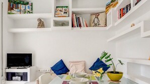 Een eethoek met een glazen tafel, witte stoelen, een televisie en boekenkasten met diverse spullen.